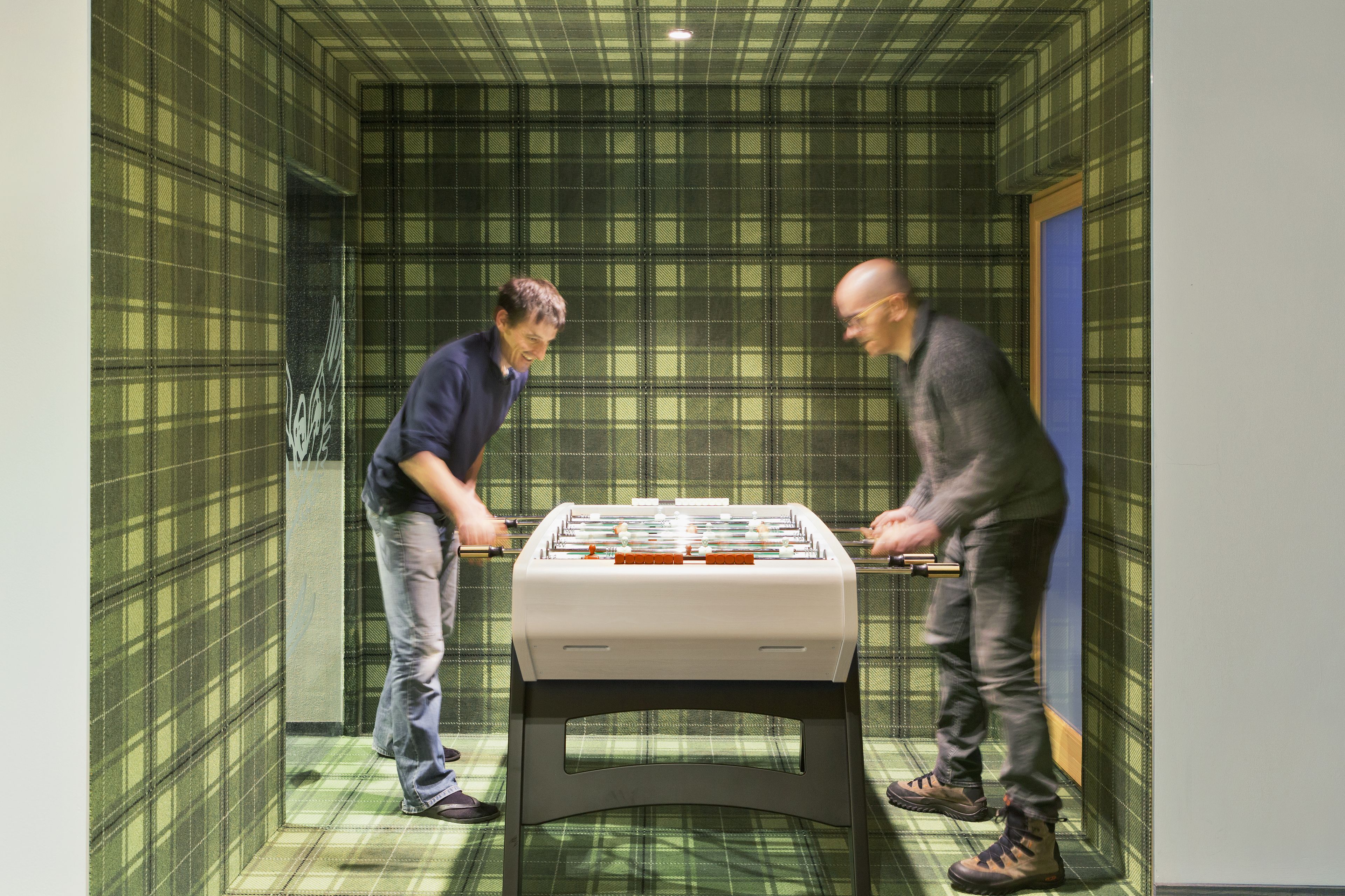 Deux hommes jouent au baby-foot dans une pièce dont un mur est décoré d’un motif à carreaux et où se trouve une table au centre.