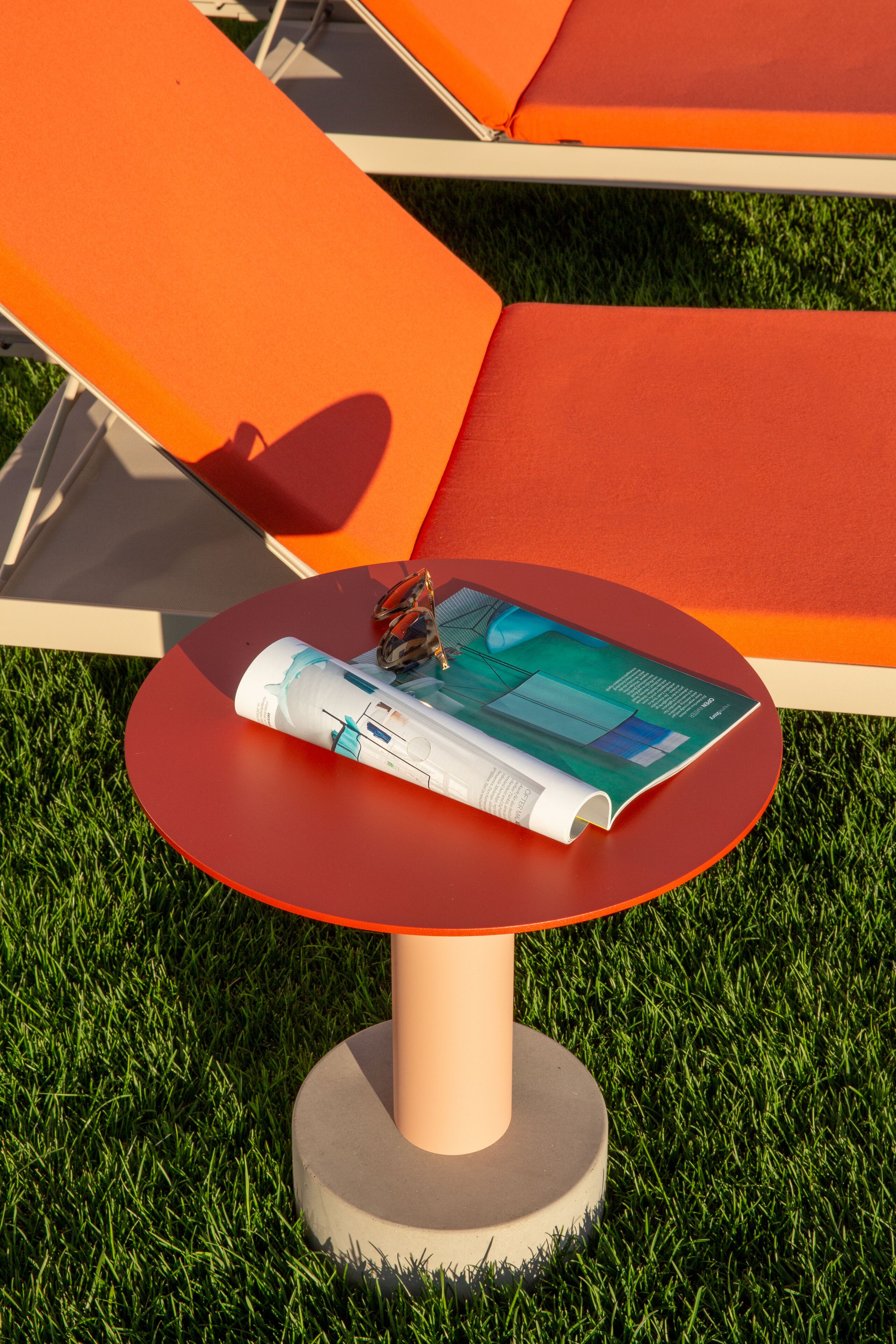 A red table with a magazine and sunglasses on it, surrounded by orange lounge chairs on a grassy area.