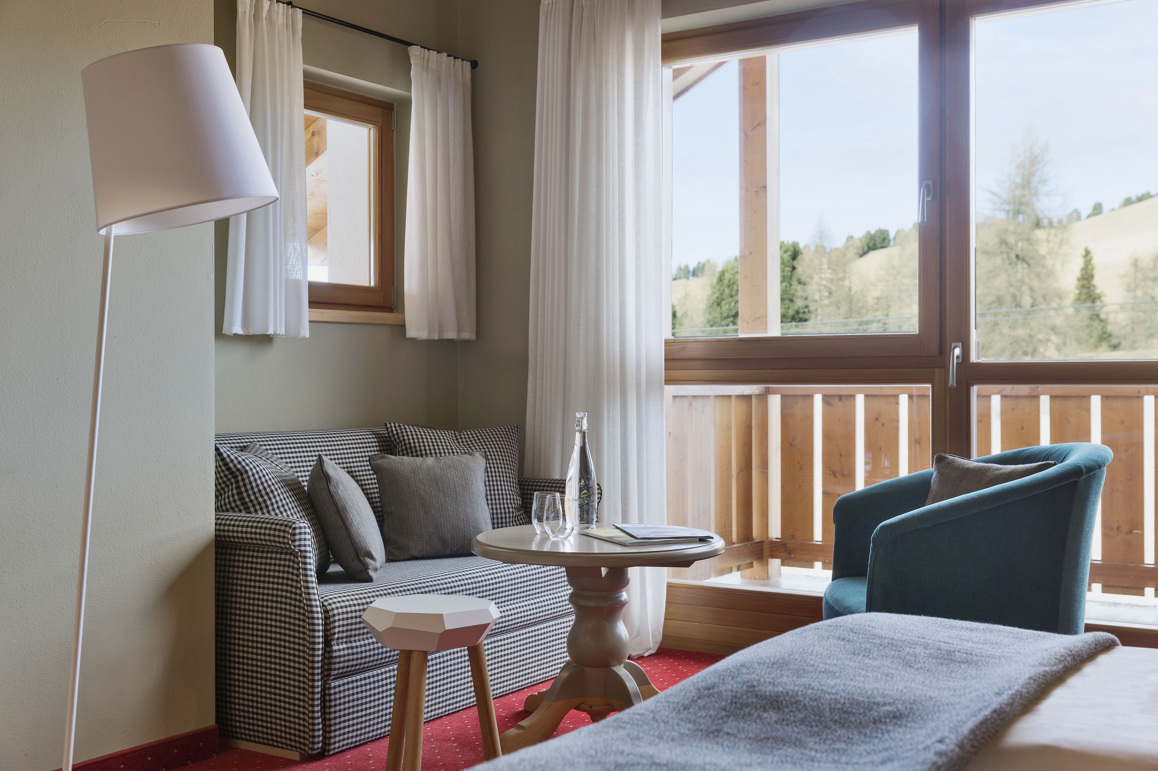 Ein Hotelzimmer mit einem karierten Sofa, einem kleinen runden Tisch mit einer Flasche und Gläsern, einem blauen Sessel und einem großen Fenster mit Blick auf Bäume.