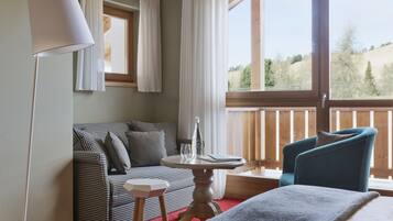 Ein Hotelzimmer mit einem karierten Sofa, einem kleinen runden Tisch mit einer Flasche und Gläsern, einem blauen Sessel und einem großen Fenster mit Blick auf Bäume.