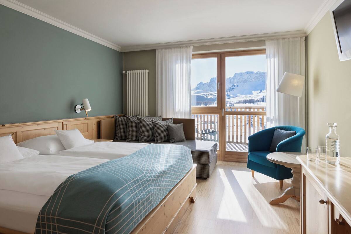 A hotel room with a large bed, a sofa, a chair, a small table, and a view of mountains through the window.