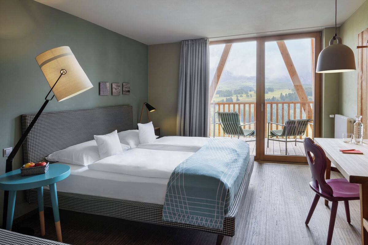 A hotel room with a large bed, a desk with a chair, a table, a balcony with chairs, and a view of the countryside.