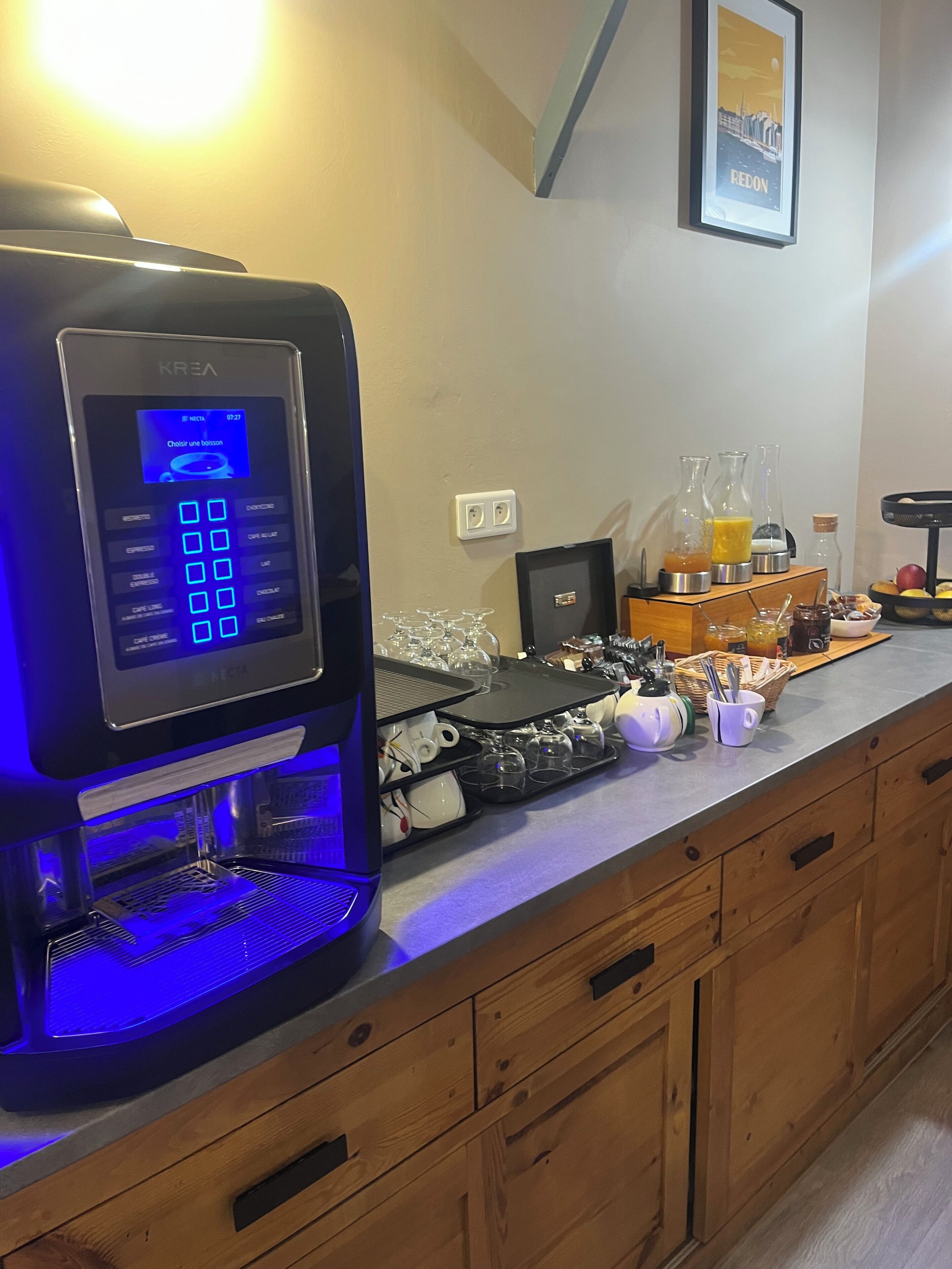 Una máquina de café KREA con pantalla digital, una bandeja de vasos y una barra de desayuno con condimentos y un cuadro enmarcado de Redon.