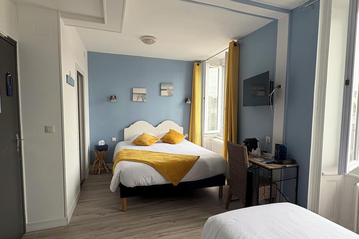 Ein Hotelzimmer mit einem Bett, einem Schreibtisch und einem Stuhl. Es gibt ein Fenster mit Vorhängen, eine Tür und einen kleinen Tisch mit einer Lampe.