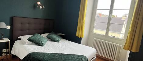 A bedroom with a large bed, a nightstand, a wall-mounted lamp, and a window with curtains.
