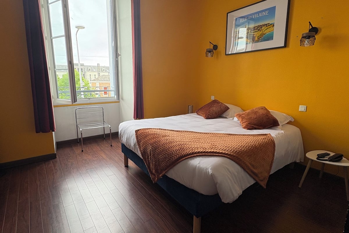 Ein Schlafzimmer mit Bett, einem Fenster mit Vorhängen, einer Wandlampe und einem Bilderrahmen.