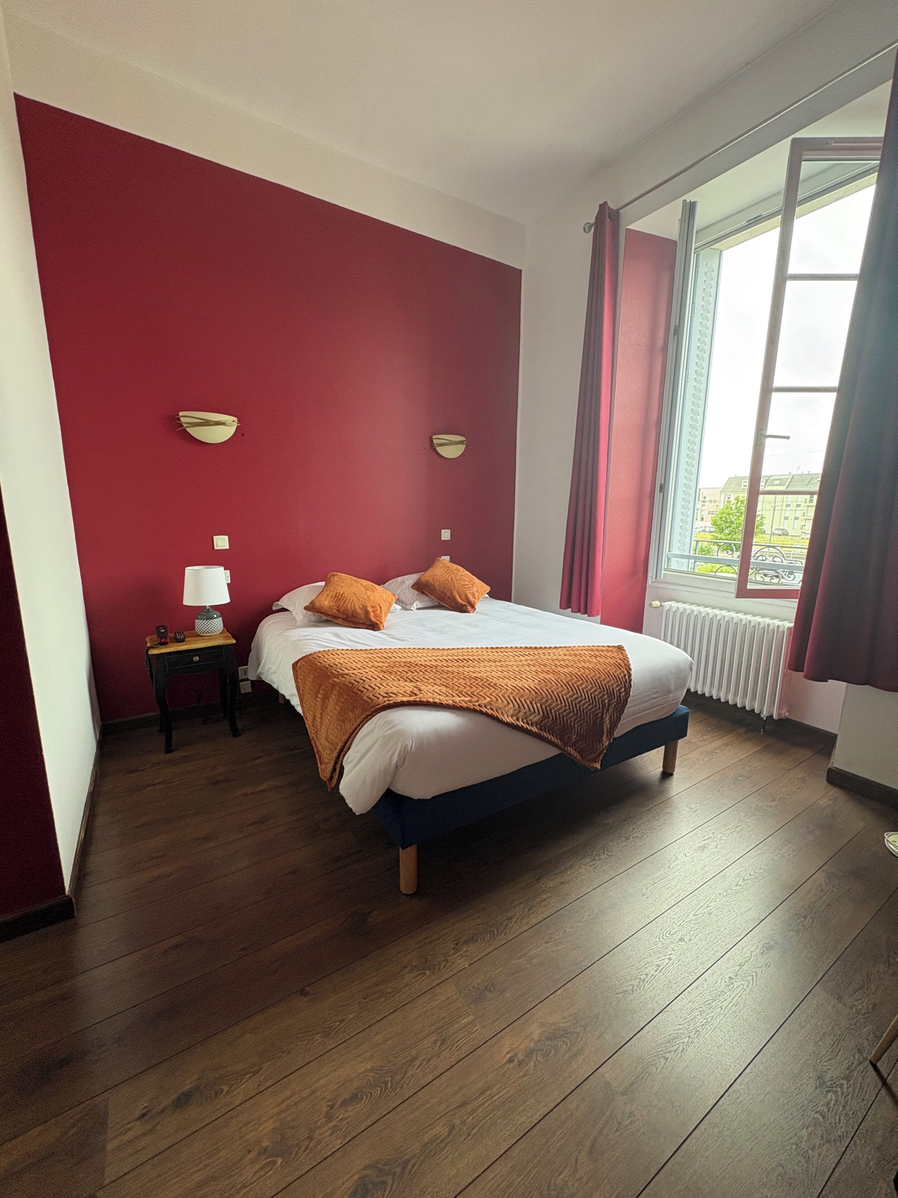 A bedroom with a wooden floor, a bed with a white mattress and orange bedding, a nightstand with a lamp, and a window with curtains.