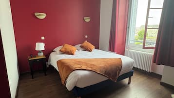 Un dormitorio con piso de madera, una cama con colchón blanco y ropa de cama naranja, una mesita de noche con lámpara, y una ventana con cortinas.