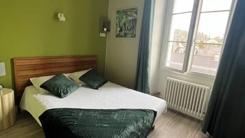 A hotel room with a wooden bed, green walls, a window with curtains, a radiator, and a small wall-mounted shelf.