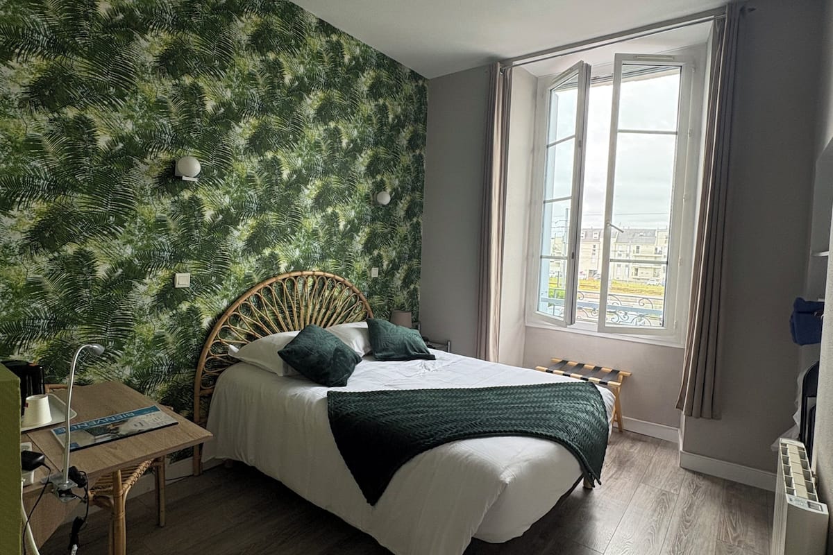 Ein Schlafzimmer mit einem Bett, einem Schreibtisch, einem Stuhl, einem Fenster und einer Wand mit einem Blattmuster.