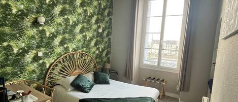 A room with a bed, a small table with a tray, a chair, a radiator, and a window with a view of buildings.