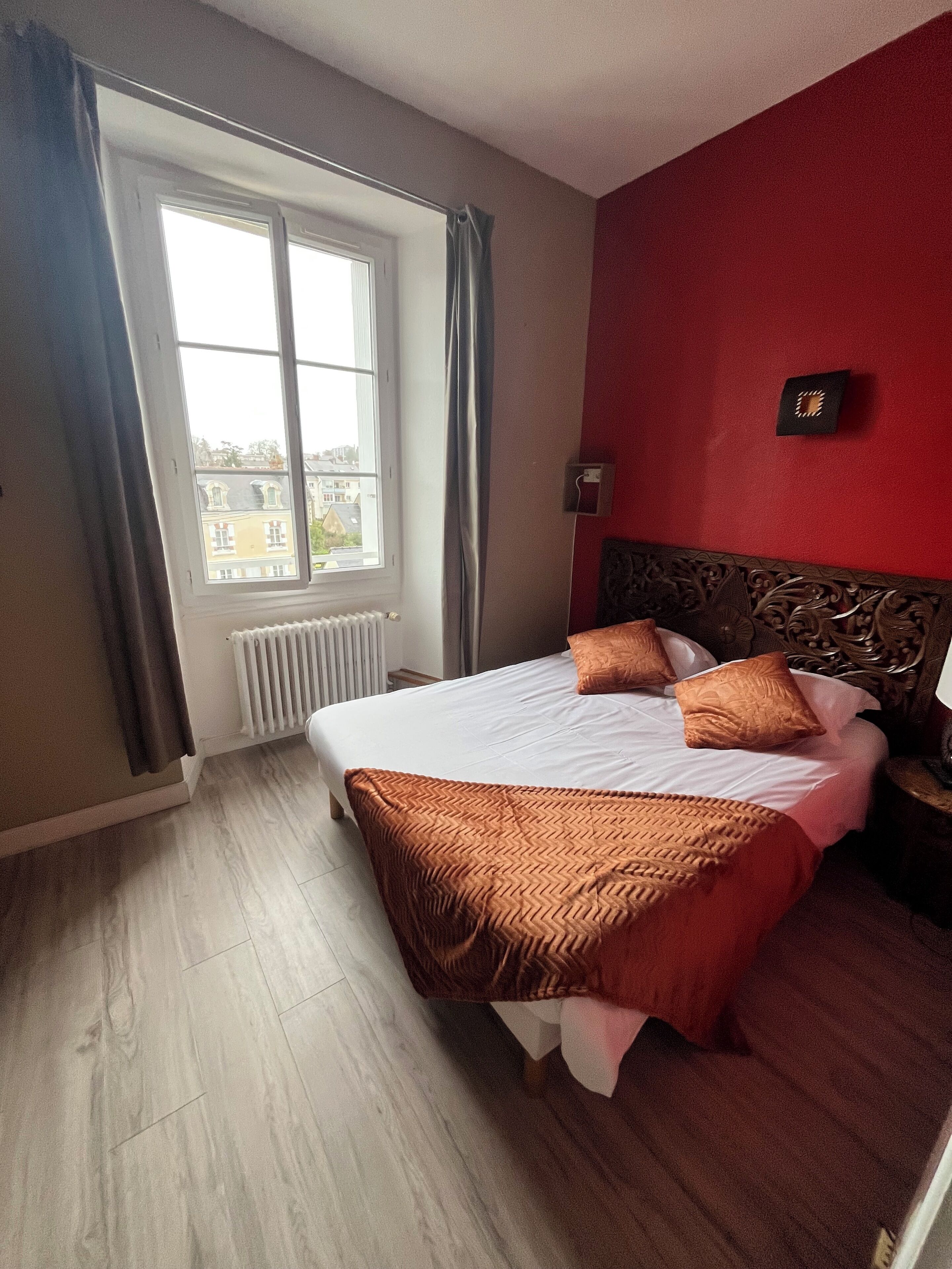 A hotel room with a bed, a window with curtains, a radiator, and a red wall.