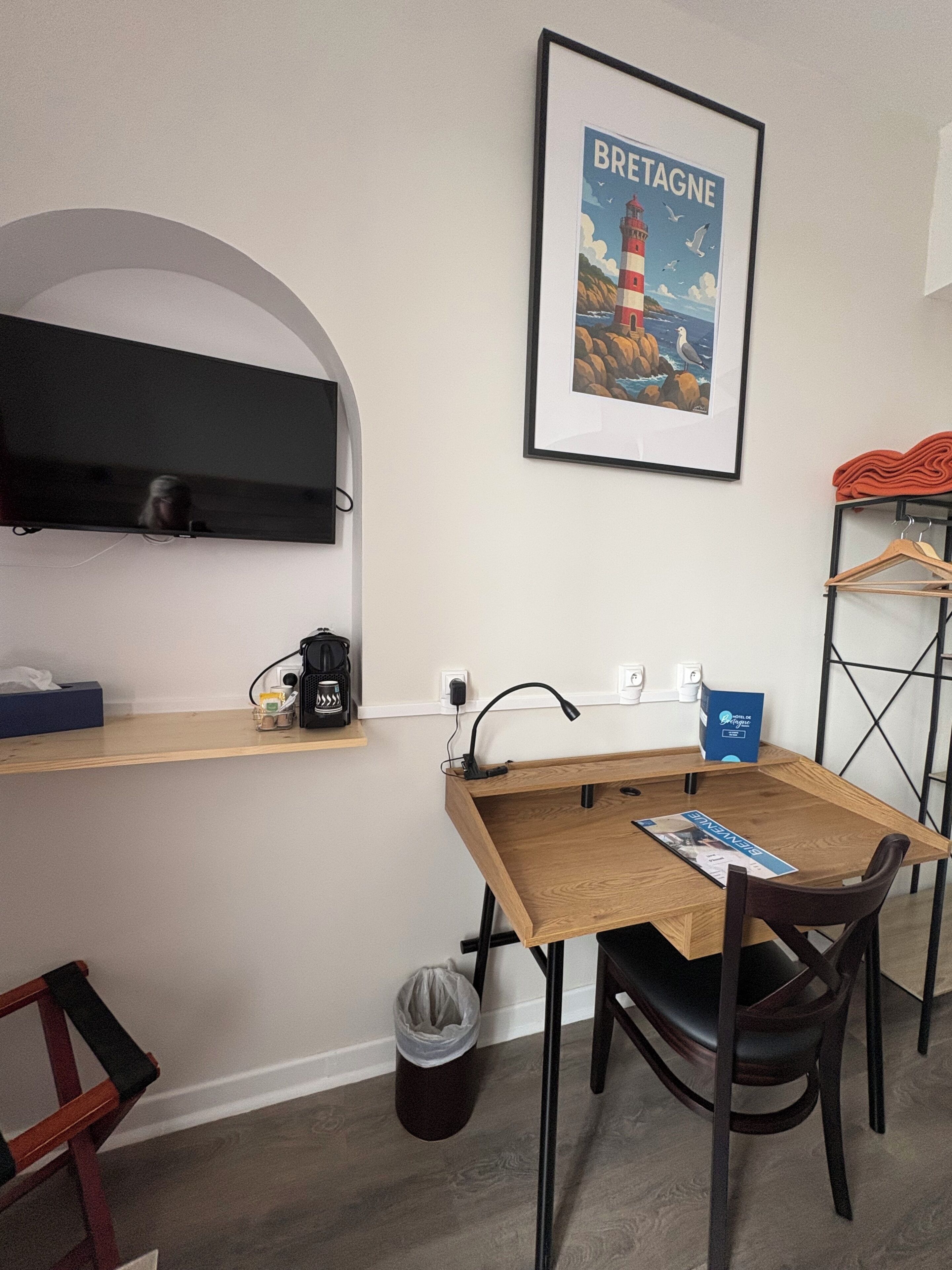 Ein Zimmer mit Schreibtisch aus Holz, einem Stuhl, einem gerahmten Bild eines Leuchtturms, einem Regal mit Kaffeemaschine und einem Kleiderhaken mit Kleidung.