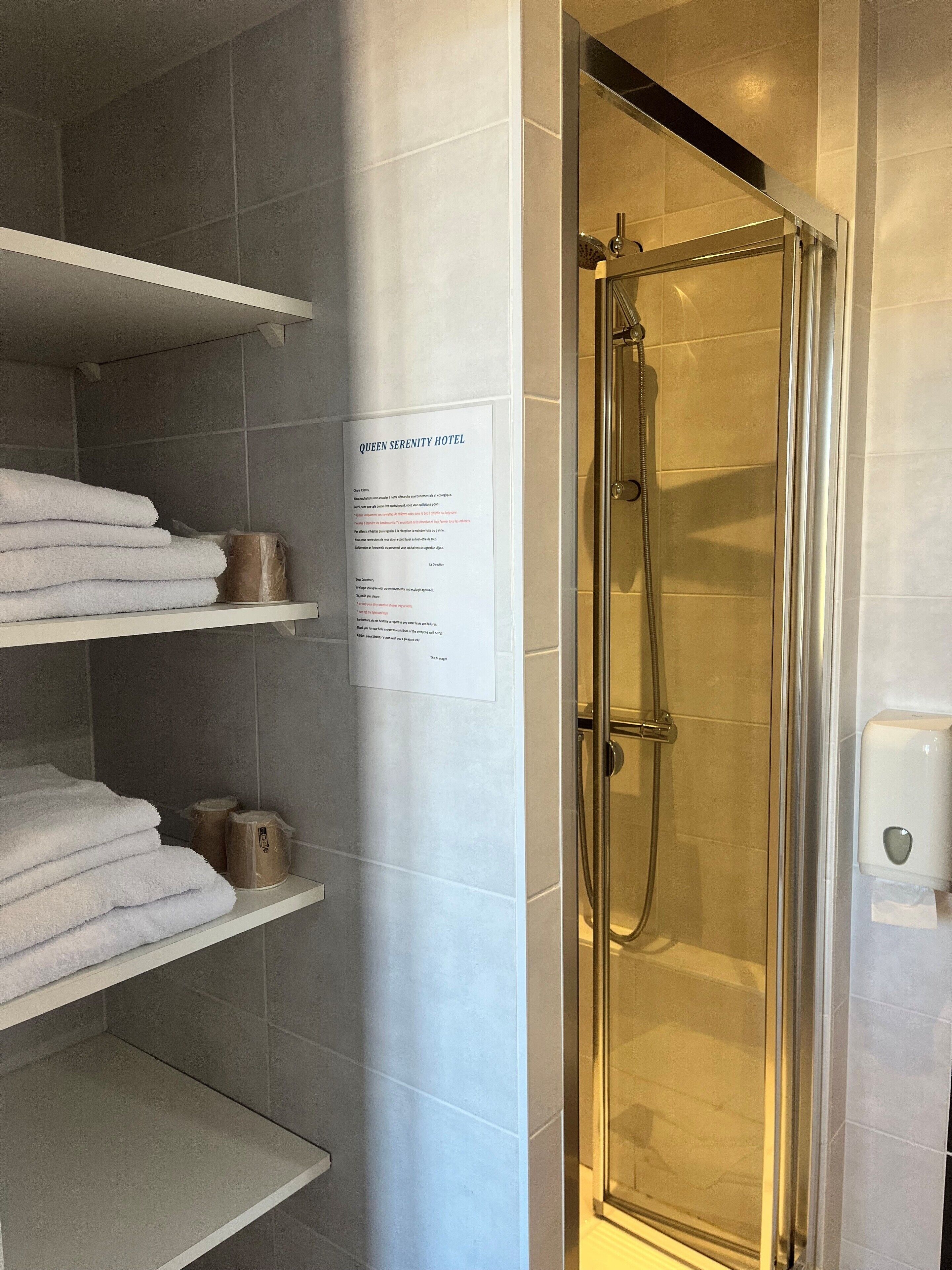 Une salle de bain d’hôtel avec une douche à parois de verre, des murs recouverts de carreaux blancs et des étagères où sont pliés les serviettes.