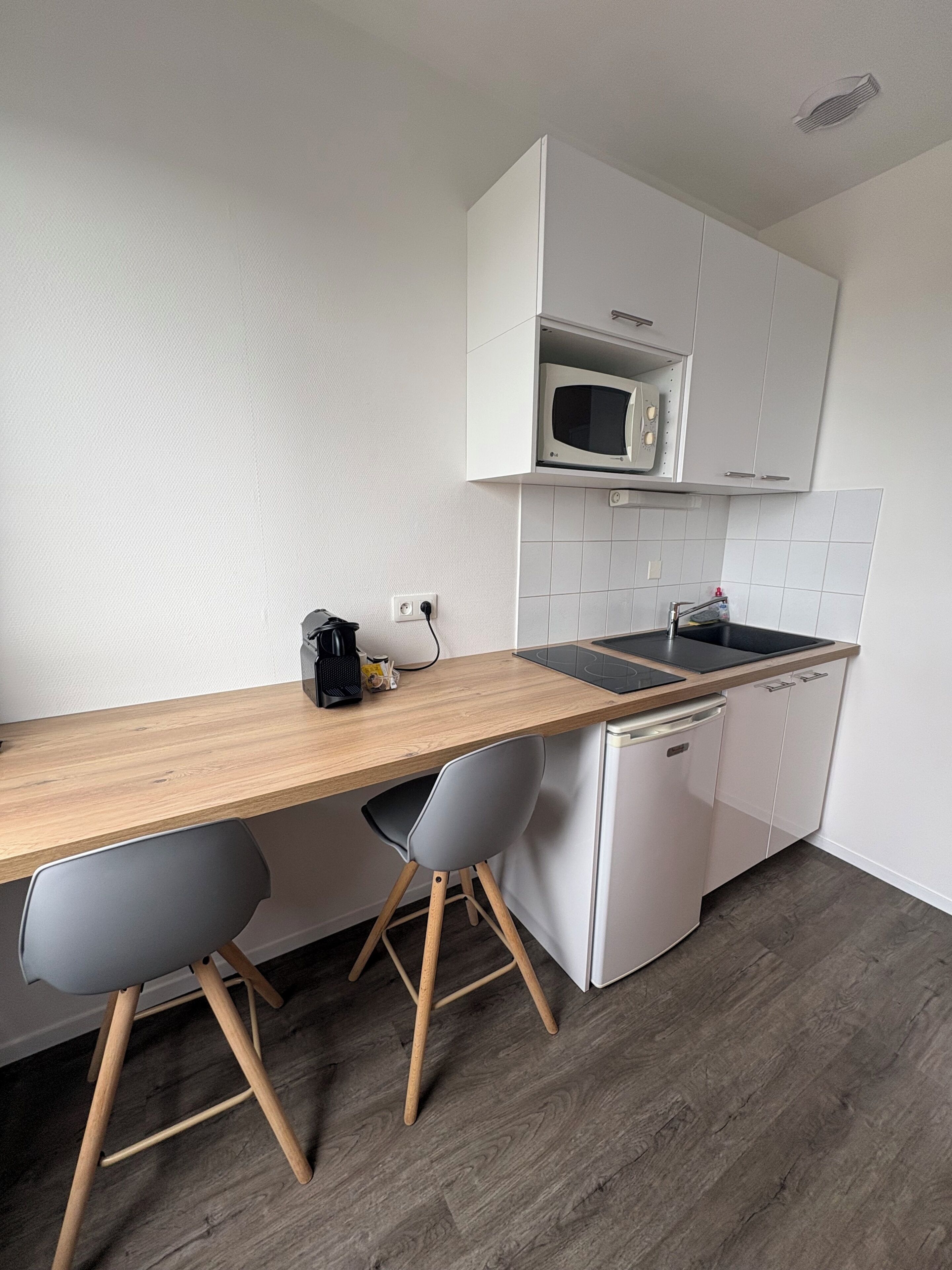 A compact kitchen with a microwave, sink, and a small dishwasher. There are two chairs and a wooden countertop.