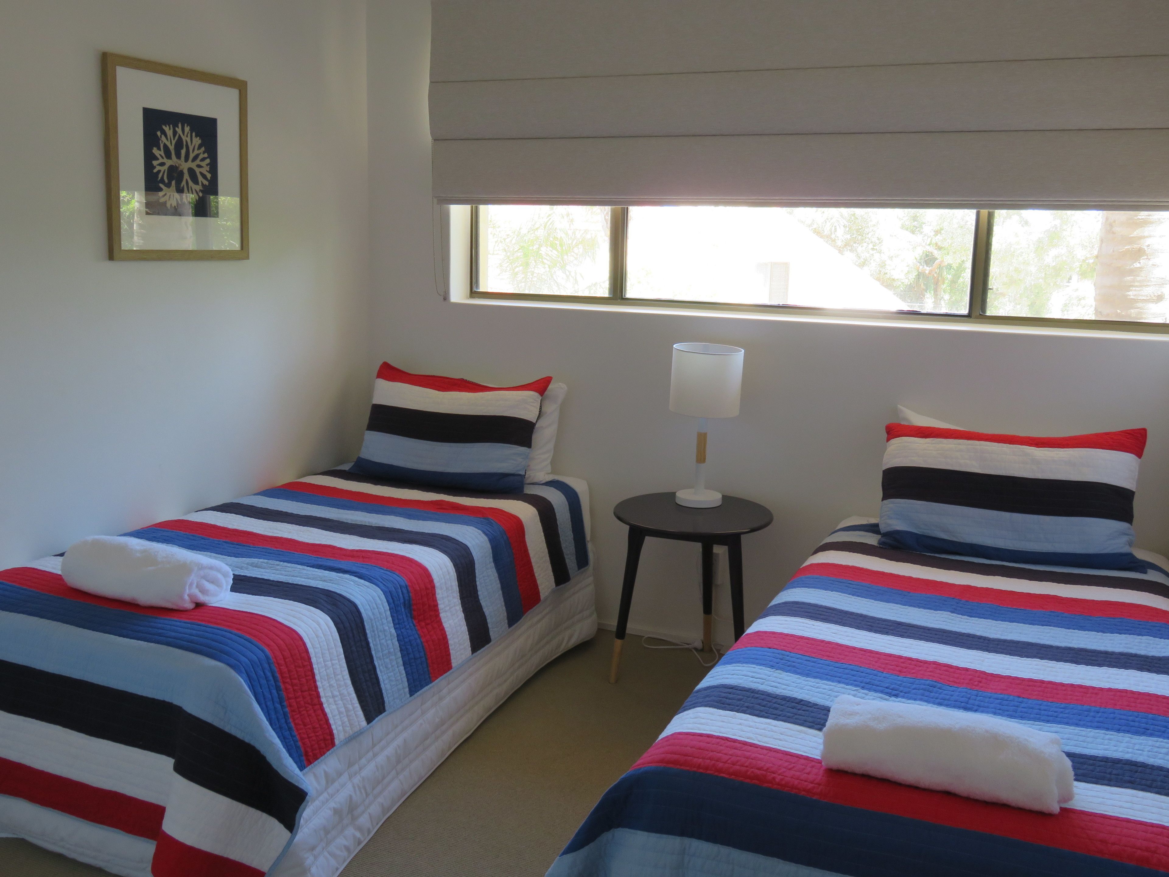 Dos camas individuales con ropa de cama a rayas, una mesita con lámpara, un cuadro en la pared y una ventana con persianas.