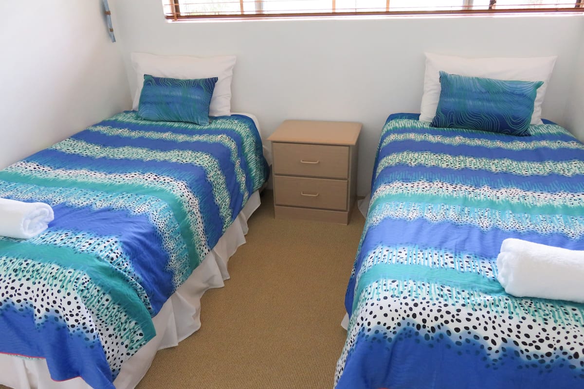 A room with two beds, each with blue and white bedding, a small bedside table, and a window with blinds.
