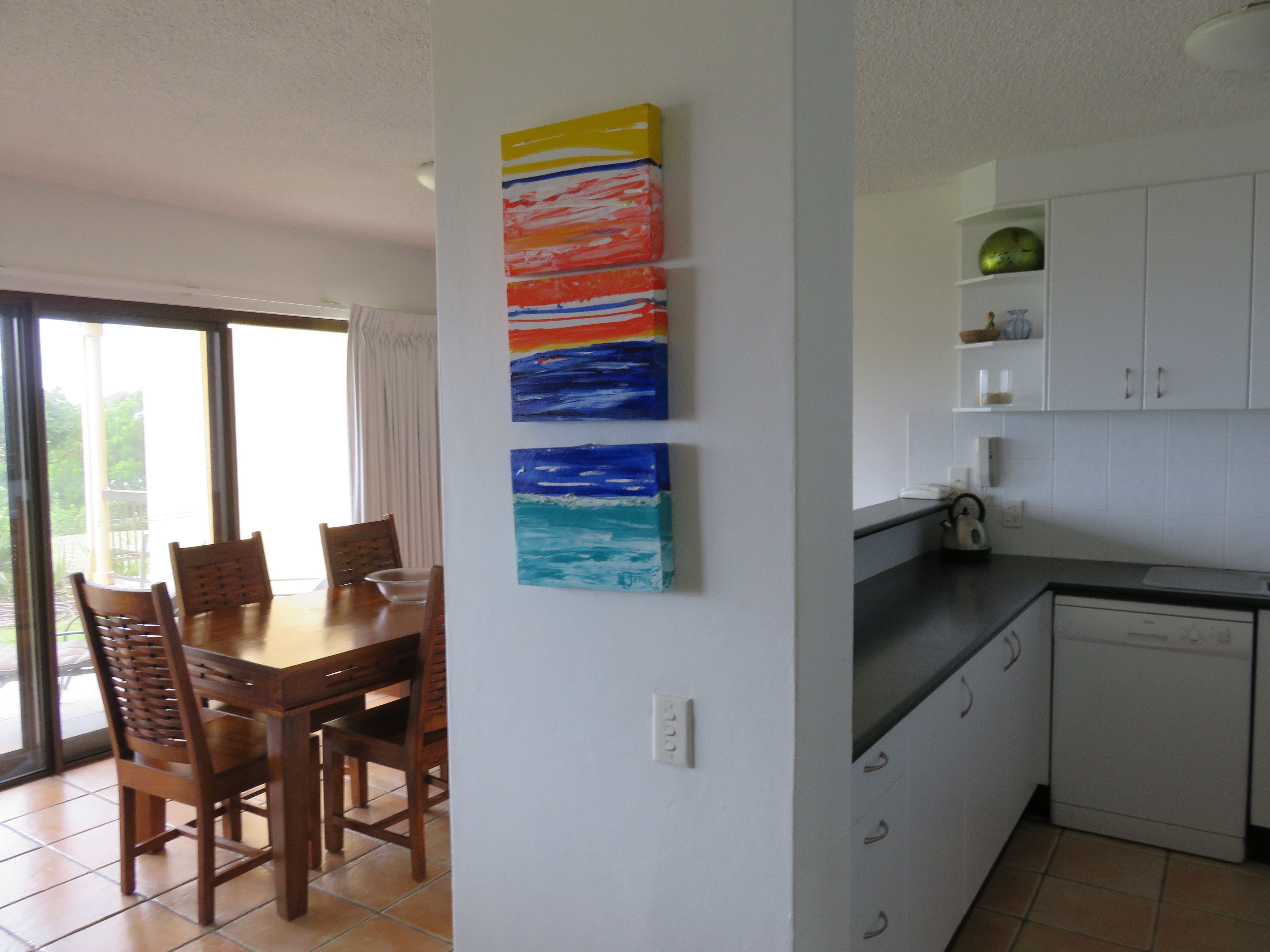 A dining area with a table and chairs, a kitchen with white cabinets, and a colorful abstract painting on the wall.