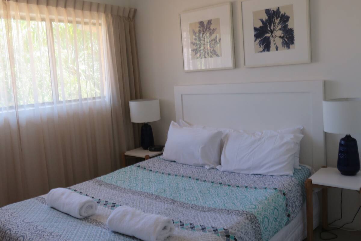 A hotel room with a bed, two bedside lamps, a nightstand, a window with curtains, and two framed wall art pieces.