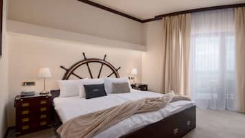 Een hotelkamer met een groot bed, een houten hoofdbord, twee nachtkastjes met lampen, een dressoir en uitzicht op het omliggende landschap door dunne gordijnen.