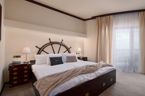Een hotelkamer met een groot bed, een houten hoofdbord, twee nachtkastjes met lampen, een dressoir en uitzicht op het omliggende landschap door dunne gordijnen.