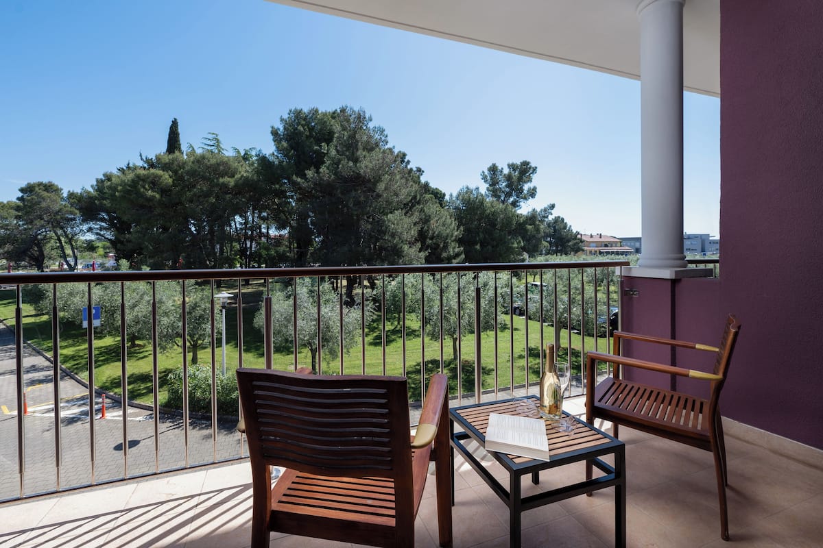 A balcony with two wooden chairs and a table, overlooking a green park and a clear blue sky.