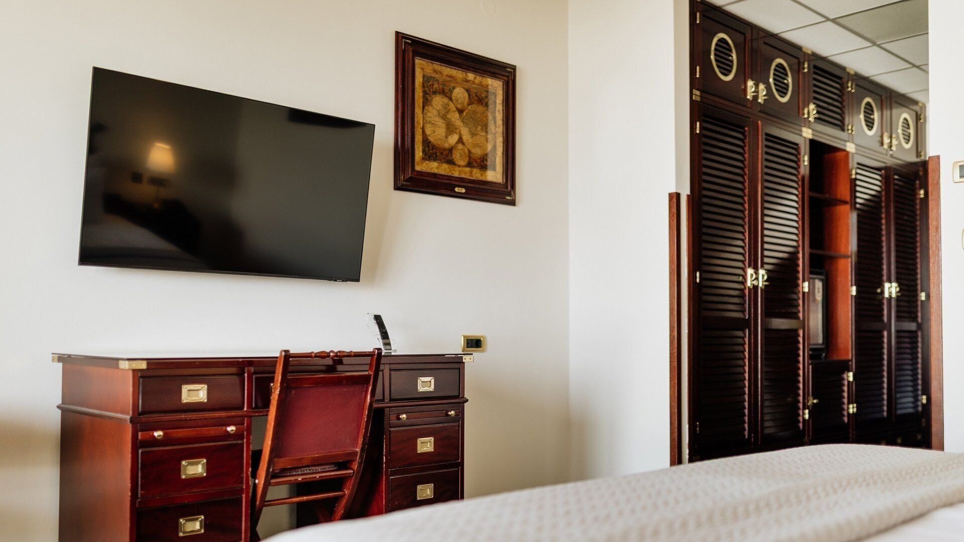 Habitación de hotel con cama, escritorio con cajones, silla, televisor montado en la pared y armario con varias puertas.