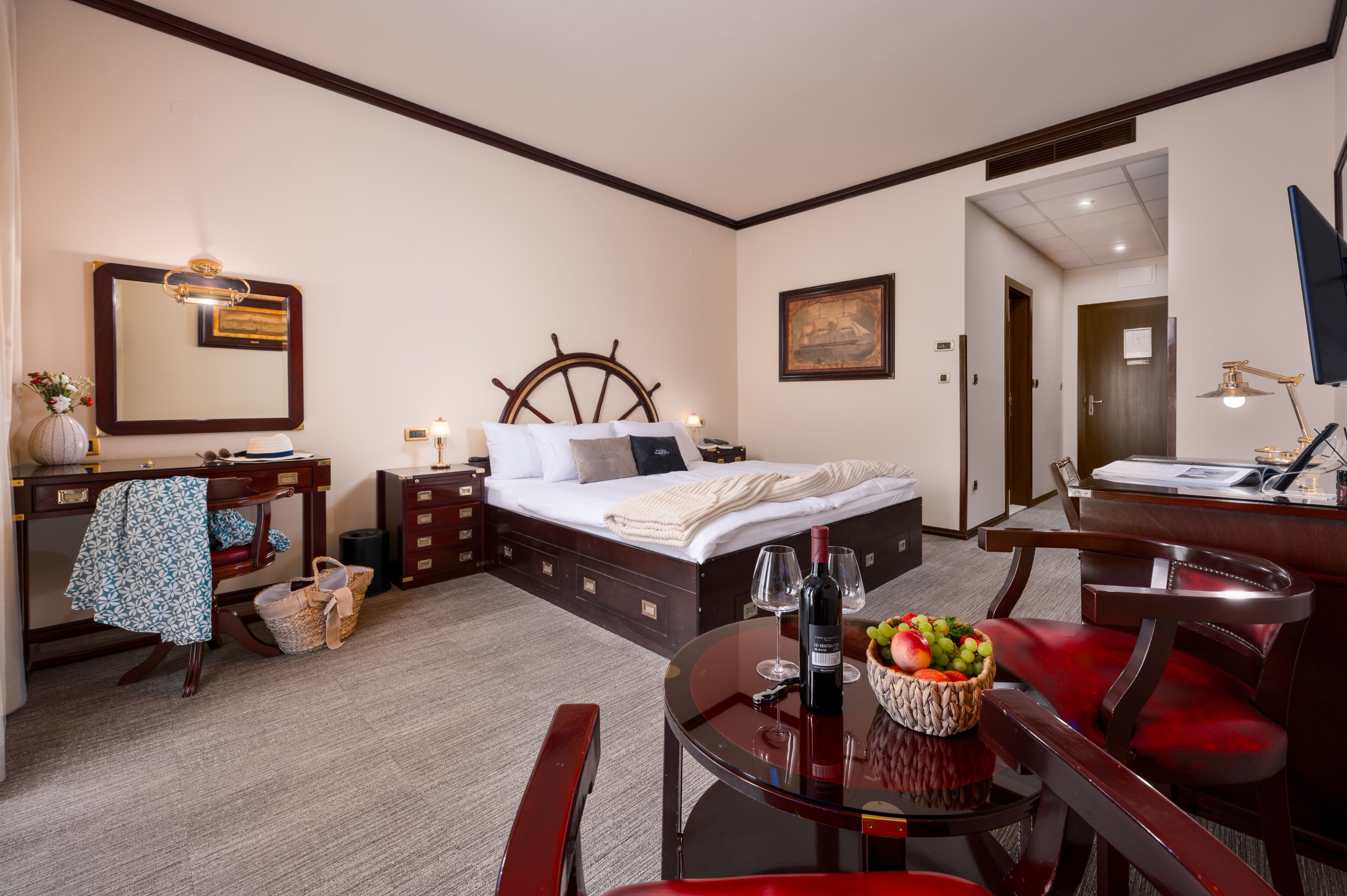 A hotel room with a bed, bedside table, desk, chair, and a dining table with a fruit bowl and wine glasses.