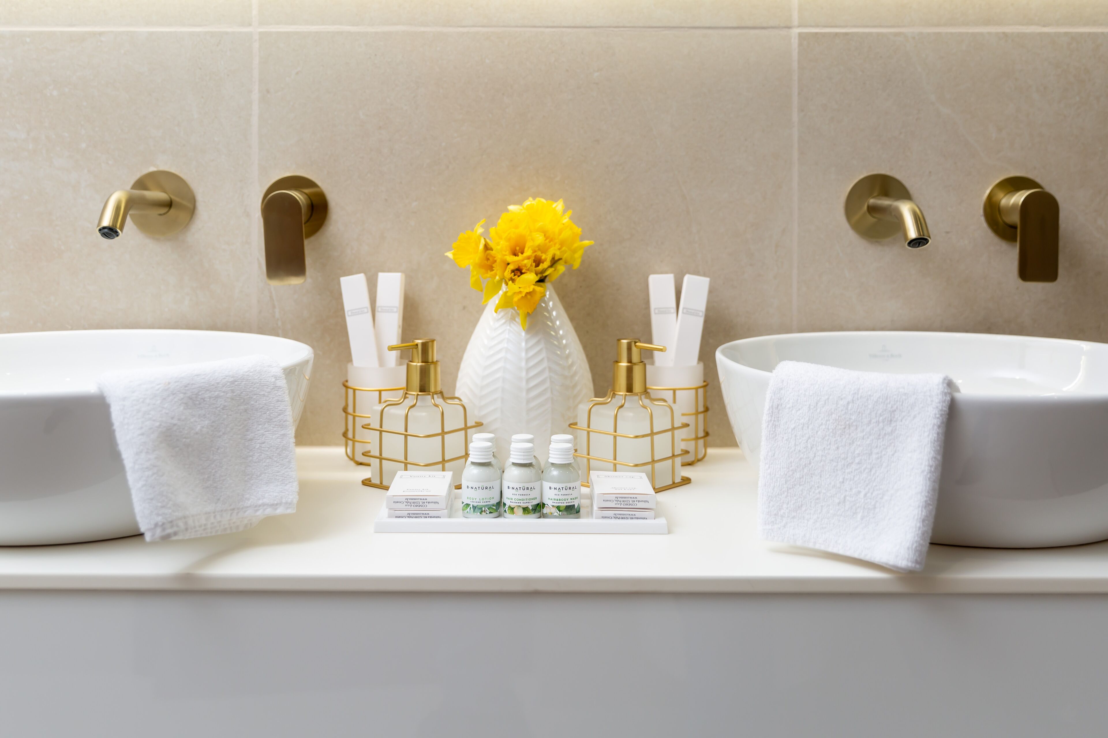 A bathroom with two sinks, a vase with yellow flowers, and toiletries on the counter.