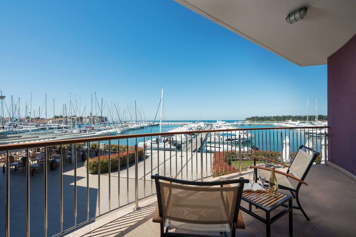 A balcony with wicker chairs, a table with a bottle and glass, and a view of a marina with boats and a bridge.