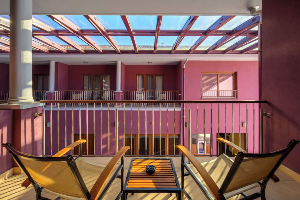 A balcony with a table and chairs, a view of the sky through a glass roof, and a pink building facade.