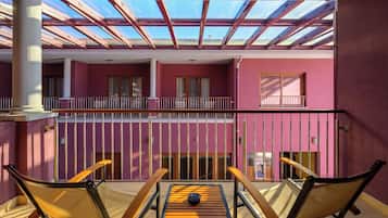 A balcony with a table and chairs, a view of the sky through a glass roof, and a pink building facade.