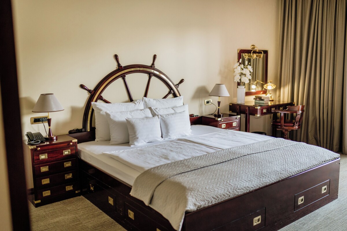 Una habitación de hotel con una cama grande, cabecero de madera, mesitas de noche, lámparas, un espejo y un aparador.