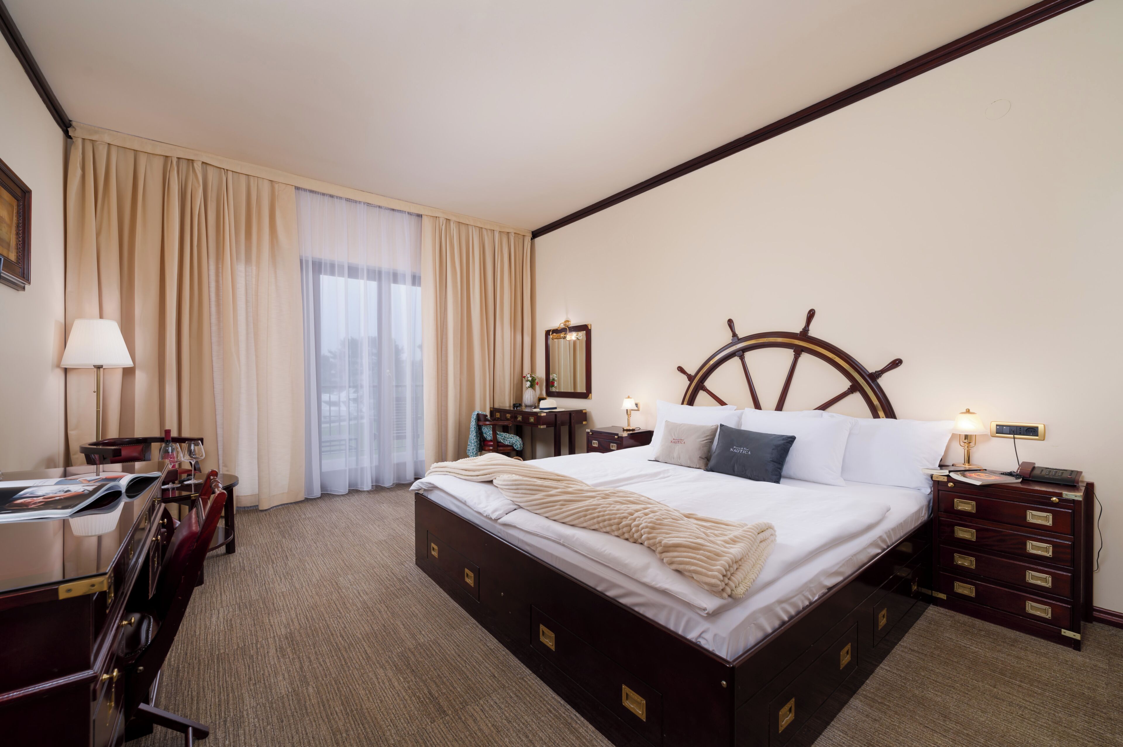 A hotel room with a large bed, a desk with a chair, a dresser, a nightstand, and a ship's wheel headboard.
