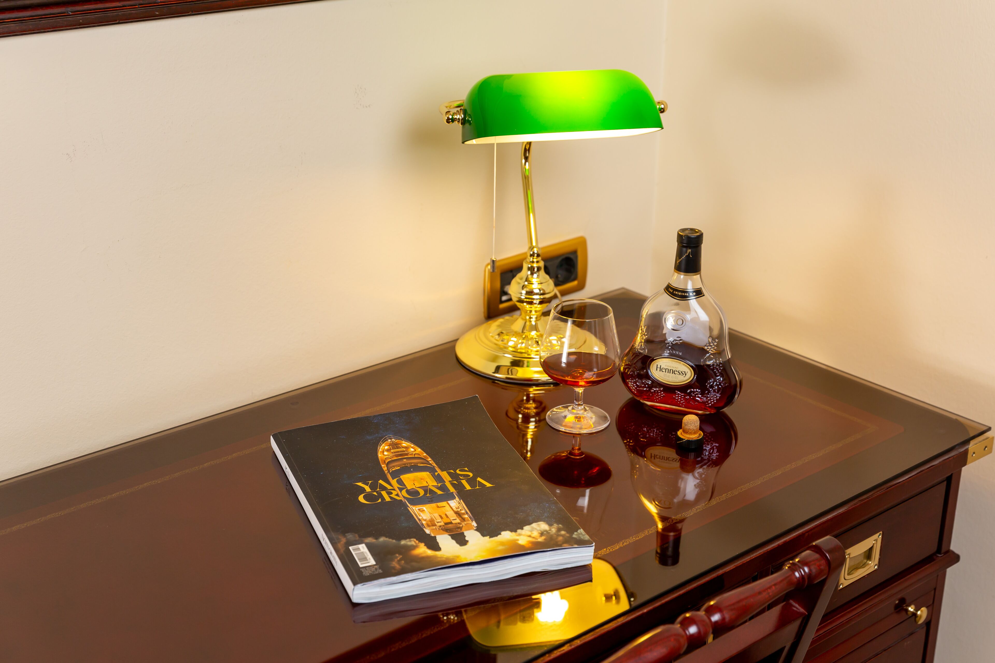 A wooden desk with a green desk lamp, a bottle of liquor, a glass, and a book titled "Yachts Croatia".