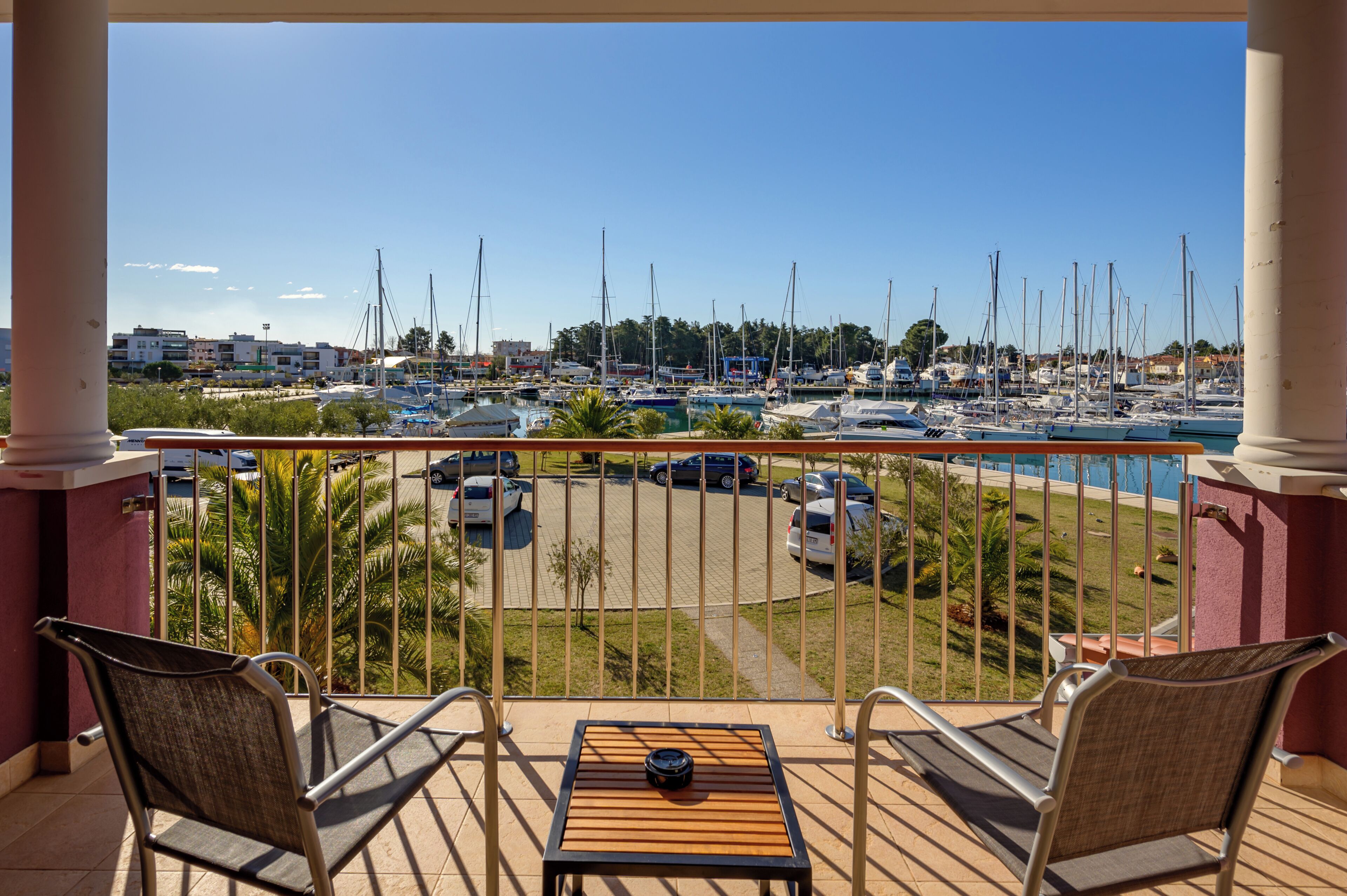 A balcony with two chairs and a table, overlooking a marina with boats and a parking area.