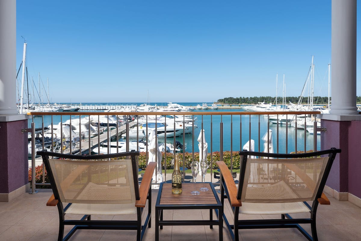 A balcony with two wicker chairs, a small table with a bottle and glasses, and a view of a marina with boats and a clear blue sky.