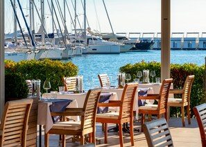 Un restaurant en bord de mer avec une terrasse, des tables dressées avec de la vaisselle et des verres, et une vue sur les bateaux au loin.