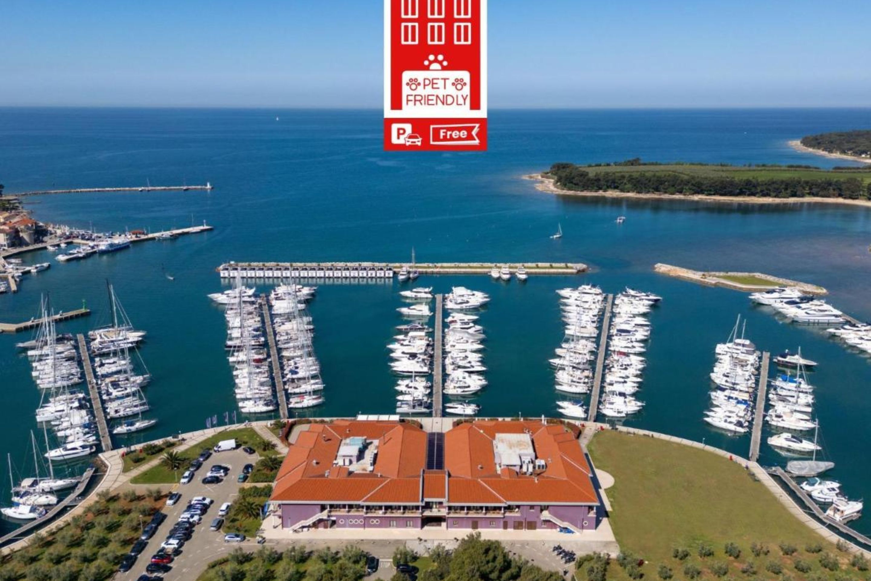 Vista aérea de un puerto deportivo con numerosas embarcaciones amarradas, un aparcamiento y un edificio con techo rojo.