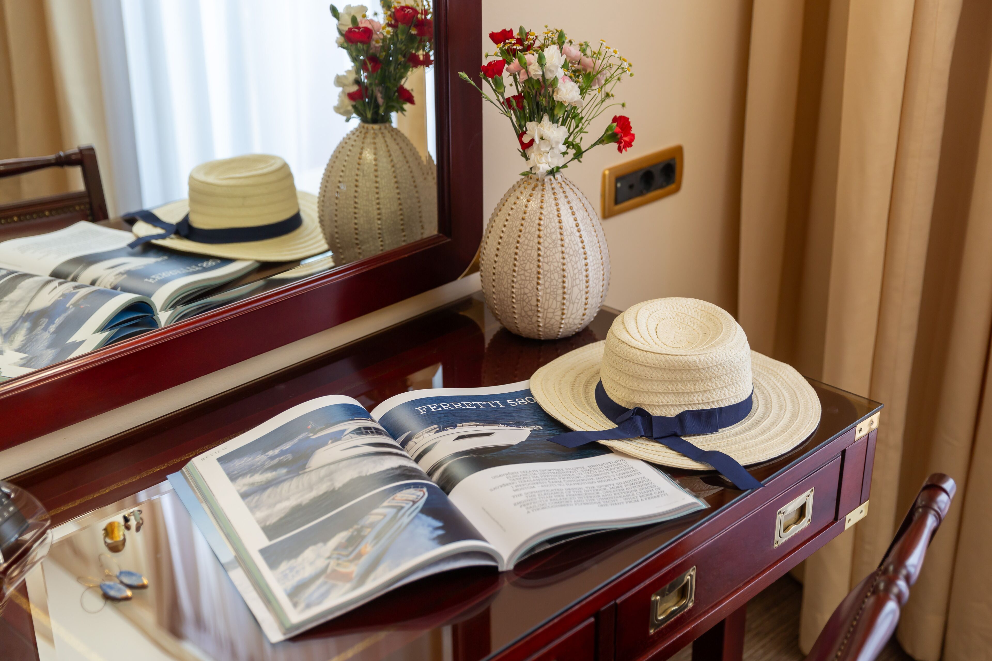 Een hotelkamer met een houten bureau, een spiegel, een bloemenvaas, een hoed en een tijdschrift.
