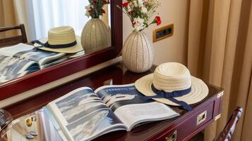 A hotel room with a wooden desk, a mirror, a vase with flowers, a hat, and a magazine.