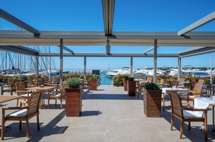 An outdoor patio with wooden chairs and tables, a view of the marina with boats, and a clear blue sky.
