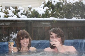 Dos personas en un jacuzzi, con arbustos cubiertos de nieve al fondo.
