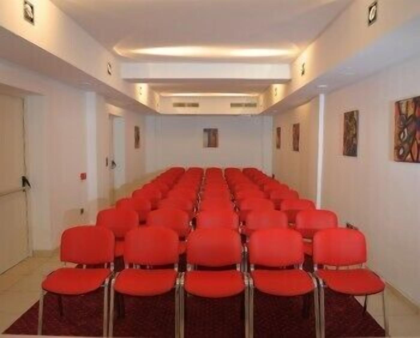 A conference room with rows of red chairs, white walls, and a carpeted floor.