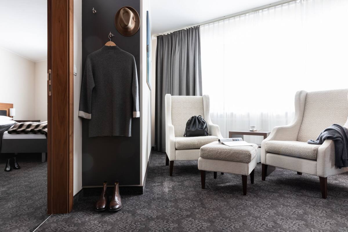 A hotel room with a bed, a wardrobe, two armchairs, a small table, and a window with curtains.