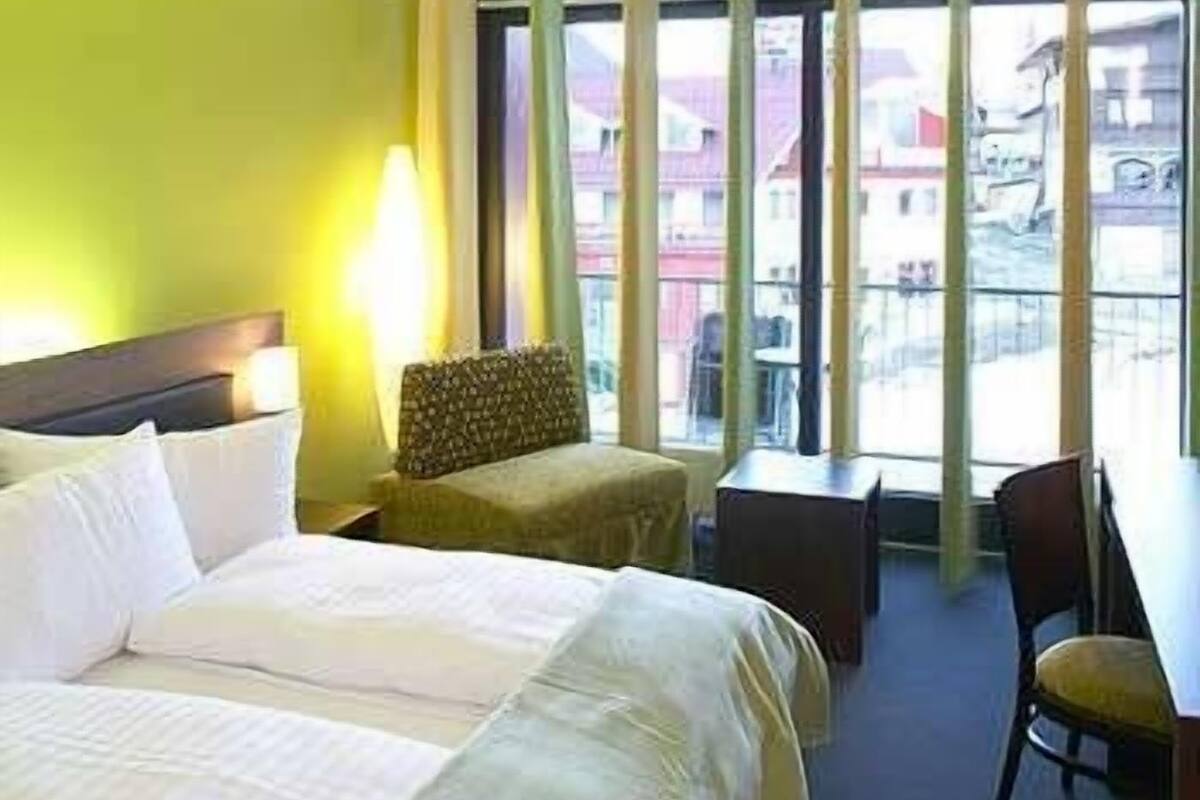 A hotel room with a bed, a chair, a small table, and a view of buildings through large windows.