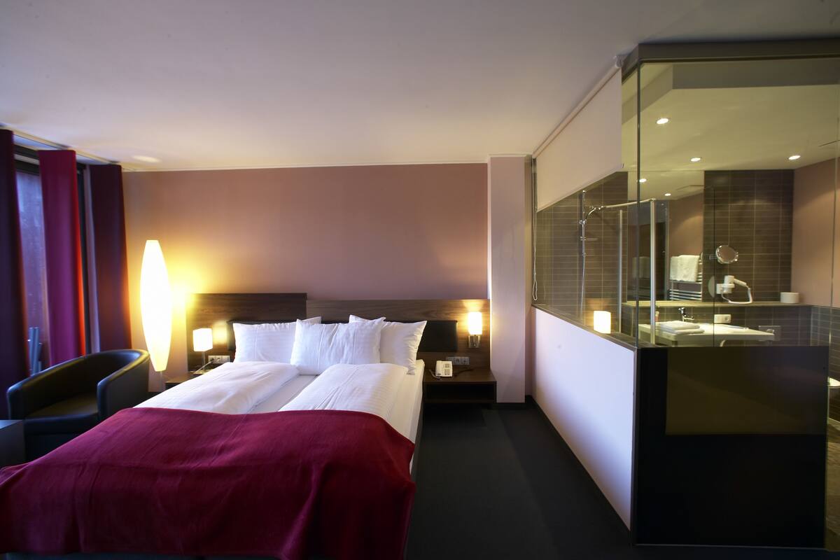 A hotel room with a bed, bedside table, lamp, chair, and a glass-enclosed bathroom.