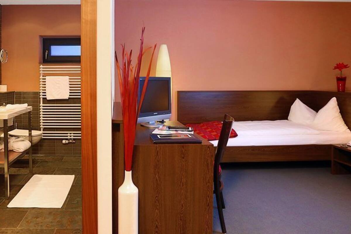 A hotel room with a bed, bedside table, and a desk with a chair. There is a bathroom visible through an open door.