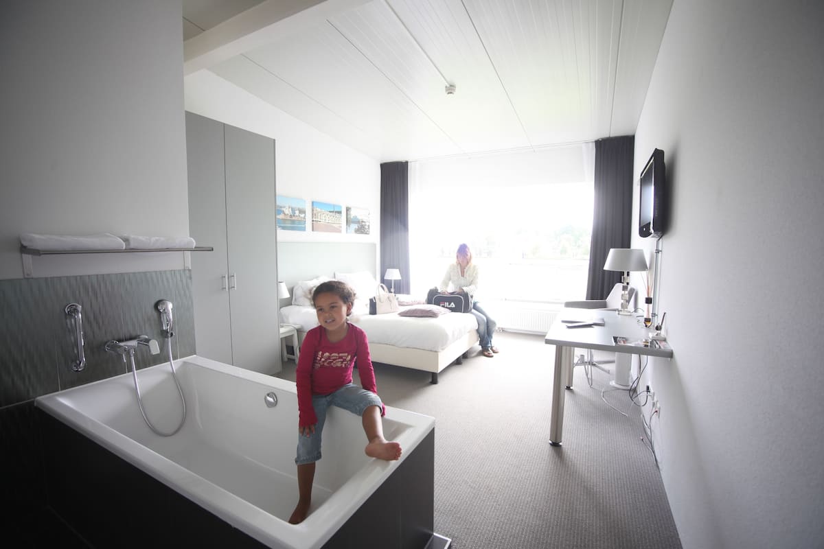 A modern hotel bathroom with a large bathtub, a separate shower area, and a seating area with a bed and a desk.