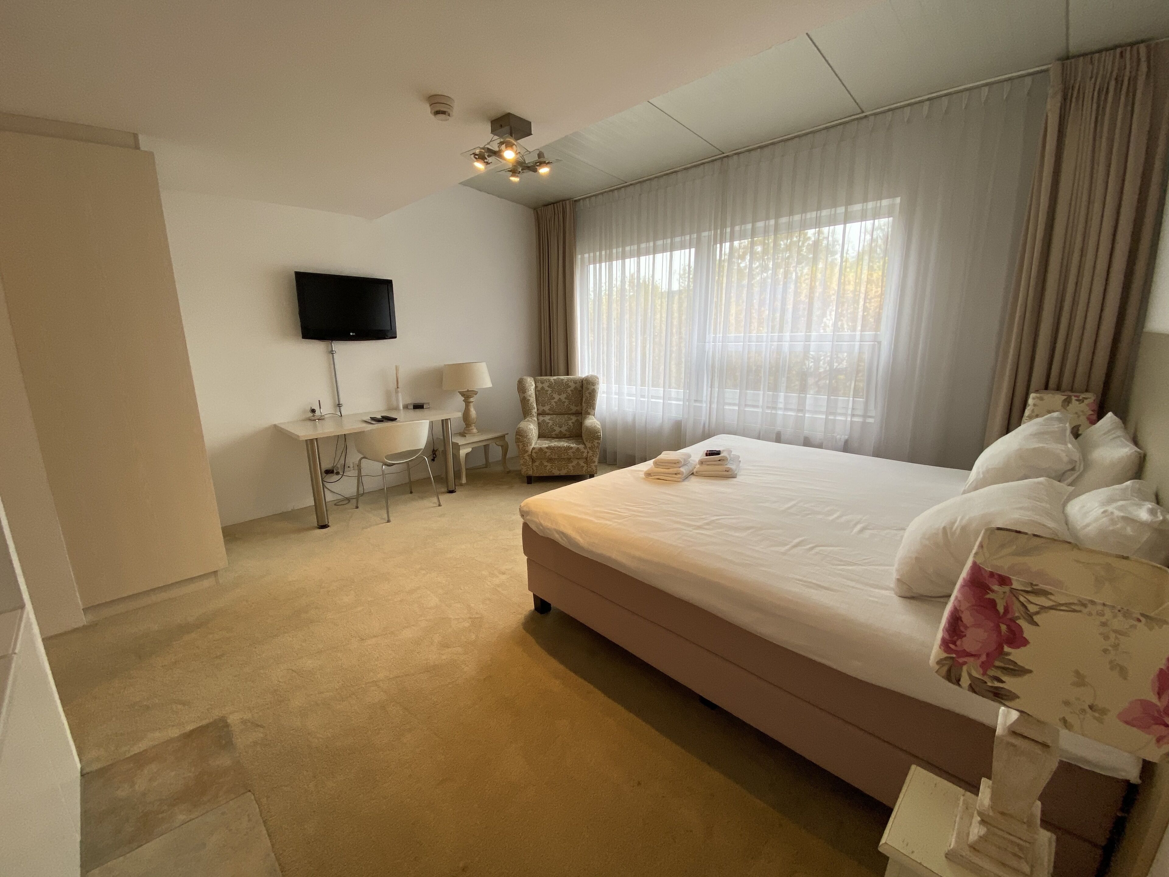 Ein Hotelzimmer mit einem großen Bett, einem Schreibtisch mit Stuhl, einem an der Wand befestigten Fernseher und einem Fenster mit durchsichtigen Vorhängen.